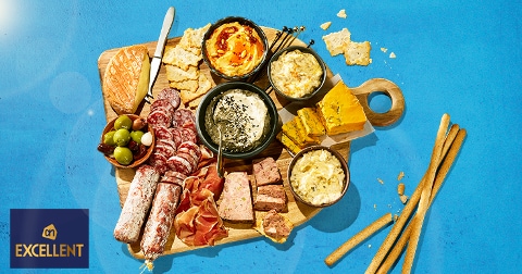 Borrelplank met verschillende charcuterie, kazen, dips, crackers, olijven en broodstengels van AH Excellent op een houten plank, gepresenteerd op een blauwe achtergrond.