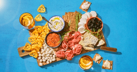 Borrelplank met verschillende hapjes zoals mini mozzarella, prosciutto, blauwe kaas, crackers, olijven, dips en drankjes met citroen op een blauwe achtergrond