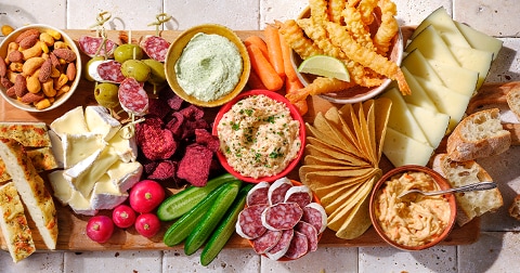 Houten plank met een variëteit aan snacks zoals kazen, charcuterie, groenten, dips, noten en crackers.