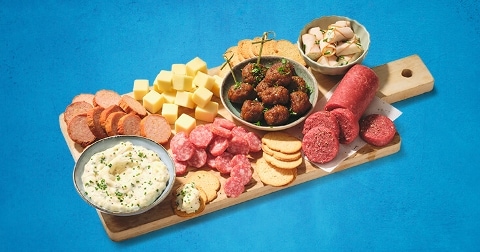 Houten serveerplank met diverse vleeswaren, kaasblokjes, gehaktballetjes met kruiden, roomkaasdip en verschillende soorten crackers.