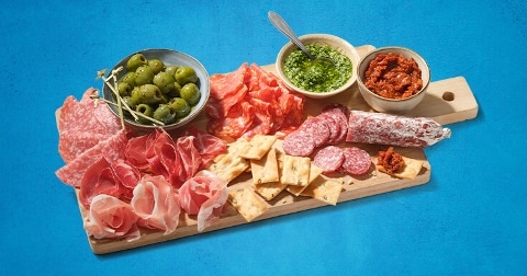 Charcuterieplank met diverse vleeswaren, olijven, pesto, tapenade en crackers op een blauwe achtergrond.