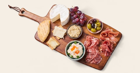 Borrelplank met verschillende soorten kazen, plakjes salami en ham, toastjes, een schaaltje hummus, een schaaltje olijven, druiven en een smeersel.