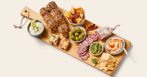 Borrelplank met vers brood, toast, droge worst, groene olijven, verschillende dips, chips en smeersels op een houten serveerplank.