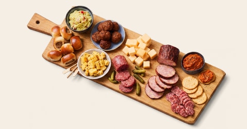 Borrelplank met verschillende hapjes, waaronder blokjes kaas, plakjes worst, mini gehaktballetjes, plakjes droge worst, rolletjes bladerdeeg, kleine augurken, dip en toastjes.
