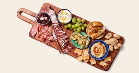 Borrelplank met diverse hapjes, waaronder plakjes salami, olijven met feta, druiven, artichok, krokante bladerdeeghapjes, hummus met pitabrood en kleine broodbolletjes.