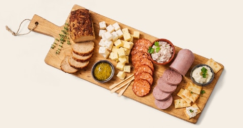 Borrelplank met gesneden brood, witte en gele kaasblokjes, verschillende soorten worst, smeersels, toast en kruiden, geserveerd op een houten plank.