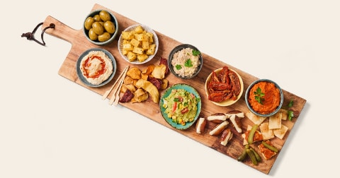 Borrelplank met verschillende mediterrane hapjes, waaronder groene olijven, blokjes kaas, hummus, chips, tonijnsalade, zongedroogde tomaten, tapenade en guacamole, geserveerd op een houten plank.