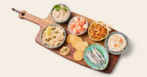 Borrelplank met diverse visdelicatessen, waaronder gerookte makreelspread, zalmblokjes, garnalen, toastjes en sardientjes op een houten plank.