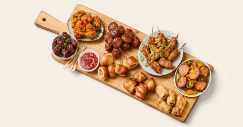 Borrelplank met verschillende snacks, waaronder kipspiesjes, gehaktballetjes, mini saucijzenbroodjes, gevuld bladerdeeg, pittige kipstukjes, worst met pepers en een bakje tomatensaus.