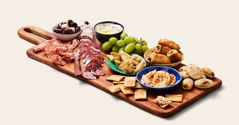 Borrelplank met verschillende tapas waaronder vleeswaren, olijven, druiven, verschillende soorten dip, crackers, gevulde bladerdeeghapjes en kleine broodjes.