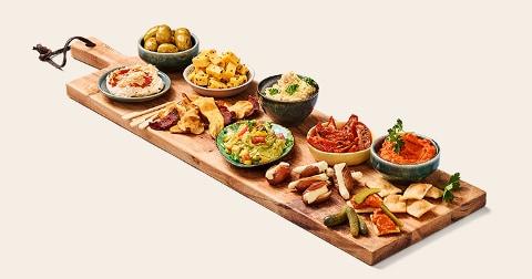Tapasplank met verschillende kleine schaaltjes gevuld met olijven, kaasblokjes, guacamole, hummus, dip, antipasti, broodstengels en stukjes platbrood.