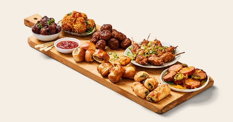 Borrelplank met verschillende hapjes, waaronder gehaktballetjes, kipspiesjes, kippenvleugels, bladerdeeghapjes, rolletjes met vulling, mini broodjes en een kommetje dipsaus.