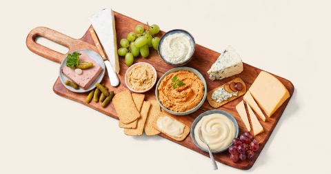 Borrelplank met verschillende soorten kaas, smeersels, paté, crackers, druiven en augurken, geserveerd op een houten plank.