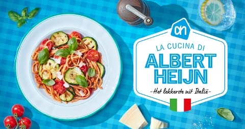 Bord met Italiaanse pasta met courgette, tomaat, basilicum en Parmezaanse kaas op een blauw geruit tafelkleed, met daarnaast het logo van Albert Heijn en de tekst "La cucina di Albert Heijn - Het lekkerste uit Italië" en een Italiaanse vlag.
