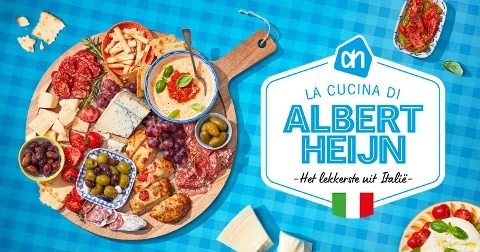 Italiaanse borrelplank met diverse kazen, vleeswaren, olijven, hummus, druiven en brood, gepresenteerd op een ronde houten plank; daarnaast een bord met Italiaanse gerechten. Promotiebeeld met de tekst: "La Cucina di Albert Heijn - Het lekkerste uit Italië".