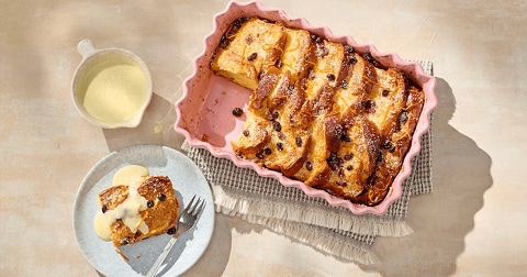 Broodpudding met rozijnen in een ovenschaal met een portie op een bord, geserveerd met vanillesaus