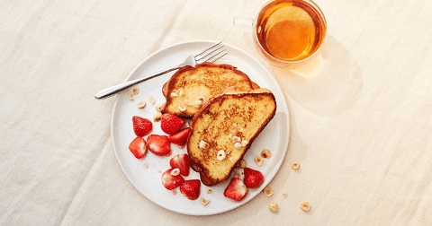 Twee sneetjes wentelteefjes met aardbeien en hazelnoten op een wit bord, met een kop thee erbij