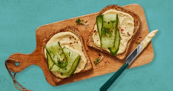Twee sneetjes volkorenbrood met hummus, komkommerschijfjes en kruiden op een houten plank met een mes