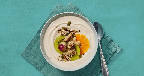 Kom yoghurt met muesli, kiwi, mandarijnpartjes, druif en noten op een turquoise servet, met lepel ernaast