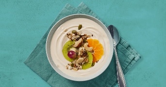 Kom yoghurt met muesli, kiwi, mandarijnpartjes, druif en noten op een turquoise servet, met lepel ernaast