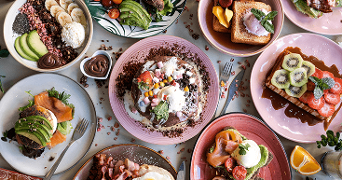 Verschillende kleurrijke ontbijtborden met onder andere pancakes, toast met fruit, yoghurt bowls met banaan en avocado, en brood met zalm, geserveerd op een tafel.