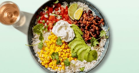 Bowl met witte rijst, avocado, mais, tomatensalsa, zwarte bonen, zure room en limoenpartjes, met een glas rosé op de achtergrond.