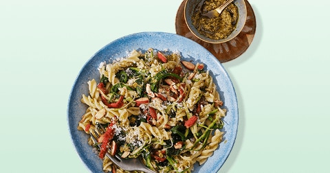 Pasta met groenten, geschaafde amandelen en pesto in een blauwe kom, met extra pesto in een schaaltje.