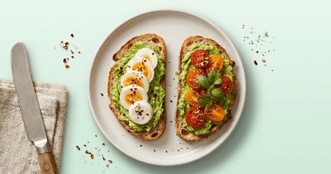 Twee sneetjes volkorenbrood met avocadospread, de ene belegd met plakjes gekookt ei en chilivlokken, de andere met gele en rode cherrytomaten en verse basilicum.