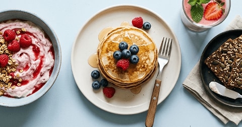 Ontbijt bestaande uit een kom yoghurt met granola en frambozen, een bord met stapel pannenkoeken en vers fruit zoals blauwe bessen en frambozen, een glas aardbeiensmoothie met munt, en een sneetje volkoren brood met zaden.