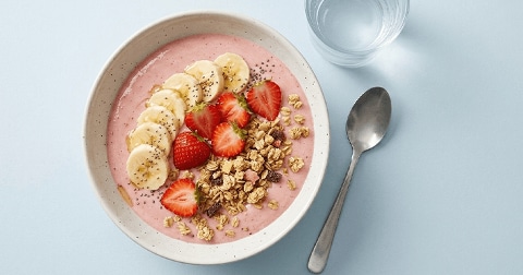Smoothiebowl met aardbeien, plakjes banaan, granola en chiazaad, geserveerd naast een glas water en een lepel.