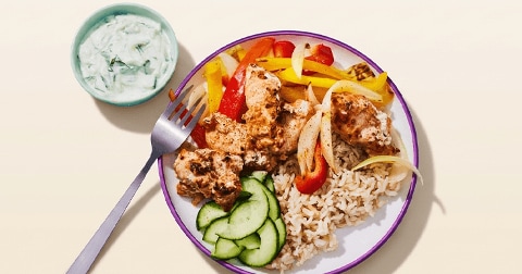 Bord met een maaltijd bestaande uit gemarineerde kip, gele en rode paprika, ui, komkommerschijfjes en zilvervliesrijst, geserveerd met een kommetje tzatziki.