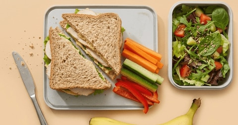 Twee volkoren sandwiches op een lunchbox met losse stukjes wortel, komkommer en rode paprika, daarnaast een bakje gemengde salade met tomaat en sesamzaadjes en een banaan.