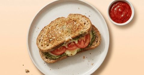 Tosti met gesmolten kaas, tomaat en pesto op een wit bord, met een bakje tomatensaus ernaast.