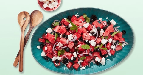 Salade van watermeloen met feta, rode ui, zwarte olijven en verse munt op een blauwgroen serveerbord, met houten saladebestek en een schaaltje feta op de achtergrond.