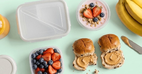 Ontbijtvoorbereiding met een bakje yoghurt met aardbeien, blauwe bessen en muesli, een bakje aardbeien en blauwe bessen, twee volkorenbroodjes met pindakaas, een tros bananen en een flesje sinaasappelsap op een lichtgroene ondergrond.