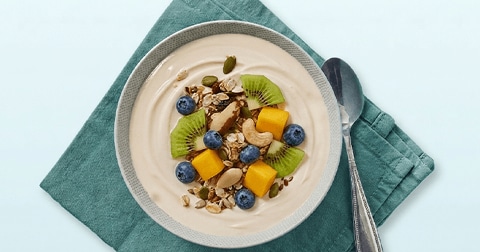 Kom ontbijt met yoghurt, muesli, blauwe bessen, stukjes kiwi, mango en cashewnoten op een groen servet.