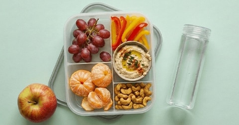 Lunchbox met druiven, mandarijnen, cashewnoten, gesneden rode en gele paprika, een bakje hummus, een appel en een flesje water op een lichtgroene ondergrond.