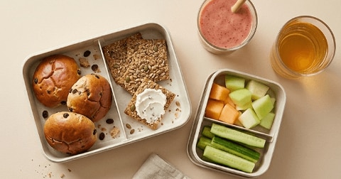 Een lunchbox gevuld met rozijnenbroodjes, volkoren crackers met zaden (een besmeerd met roomkaas), stukjes komkommer, meloen en honingmeloen, met daarnaast een glas appelsap en een glas aardbeien smoothie.