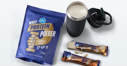 Proteïneproducten van Albert Heijn, waaronder een zak whey proteïnepoeder vanille smaak, een shaker met proteïnedrank en twee proteïnerepen, één van witte chocolade en één van karamel.