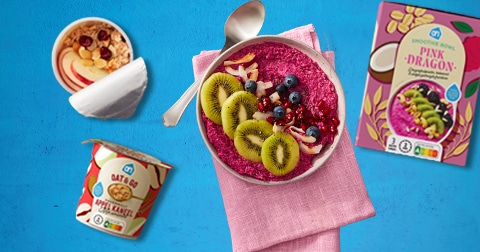 AH Pink Dragon smoothie bowl met vers fruit, een AH Oat & Go appel-kaneel cup en een bakje met yoghurt, muesli en fruit gepresenteerd op een blauwe achtergrond