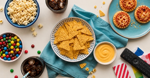 Schaal met tortillachips en kaassaus, omringd door popcorn, mini pizza’s, gekleurde M&M’s, frisdrank in glazen en een afstandsbediening, op een licht oppervlak met servetten; ideaal voor een gezellige filmavond of snackmoment.