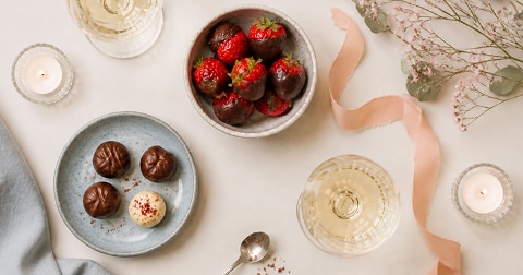 Aardbeien gedoopt in chocolade in een schaal, pralines op een blauw bord, glazen witte wijn, brandende kaarsjes en bloemen als tafelversiering.