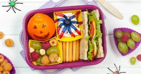 Lunchbox met Halloween-thema met mandarijn als jack-o'-lantern, crackers met groentesticks, toast met sla en tomaat, fruit en snackgroenten, omringd door decoratieve spinnen.
