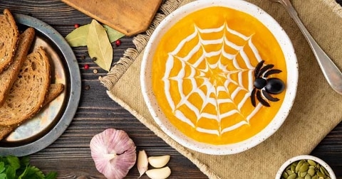 Pompoensoep met een decoratief spinnenweb van room en een olijfspin, geserveerd met brood, knoflook en pompoenpitten