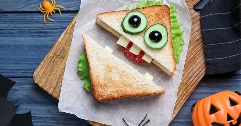 Tosti in de vorm van een monster met komkommer en olijf als ogen, kaas als tanden en sla op een houten plank, versierd voor Halloween