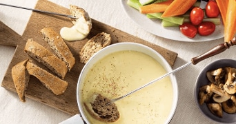 Pan met gesmolten kaasfondue met stukjes stokbrood op een houten plank, gesneden groente zoals wortel en bleekselderij, cherrytomaten en een bakje gebakken champignons