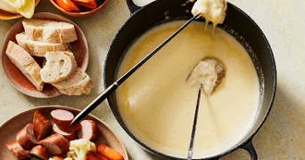 Kaasfondue in een pan met dipstokjes, geserveerd met stukjes brood, worst, bloemkool en wortel op borden