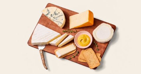 Assortiment van verschillende kazen op een houten plank met toast, kruiden en een kommetje mosterd.