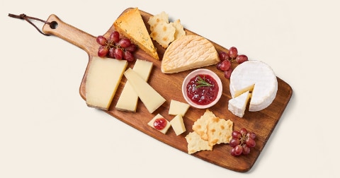 Verschillende soorten kaas op een houten borrelplank, geserveerd met druiven, crackers en een schaaltje rode jam.
