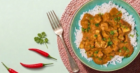 Bord met rijst en kipcurry, gegarneerd met koriander, naast een vork, rode chilipepers en een korianderblad.